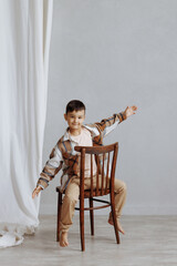 A young boy is standing in front of a wooden chair. He is wearing a plaid jacket and tan pants. The...