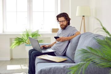 Bearded brown hair Caucasian man with laptop on knees sits on sofa with postal envelope and uses mobile phone to write review about product ordered on Internet is in living room of house