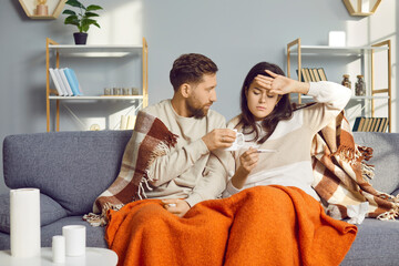 Unhealthy man and woman are sitting at home in the living room, wrapped in warm blanket and feeling flu symptoms. Young couple looks at thermometer that shows high temperature. Healthcare.