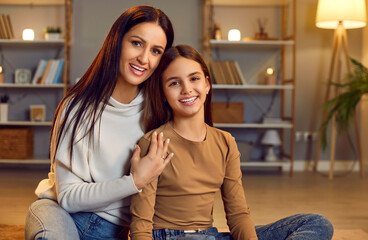 Portrait of happy mother and pre teen child at home. Happy mom and tween daughter in casual clothes sitting together on floor in living room, looking at camera and smiling. Family, love, care concept