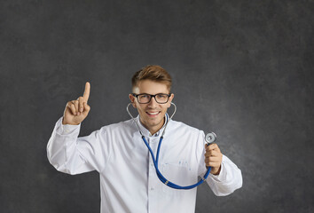 Funny smiling male doctir general practitioner shows that he has idea for your diagnosis. Glad medical worker listens to heartbeat with stethoscope and raises his finger up standing on gray background - 796951723