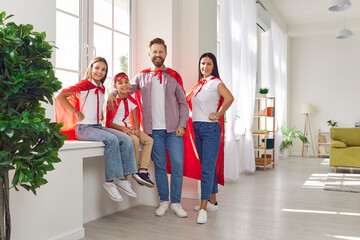 Portrait of smiling young happy family with children in superhero costumes standing near the windowsill at home and looking cheerful at camera. Love, care, family time and leisure concept.