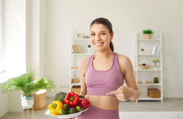 Young woman keeps fit by working out and eating healthy food. Portrait of happy beautiful girl in pink sports top smiling and pointing at plate with colorful raw vegetables. Fitness concept - 796960527