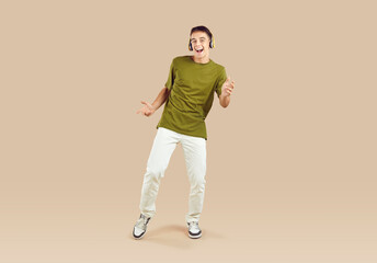 Adolescent boy dancing and singing in headphones enjoying music on beige background in studio. Full length Caucasian teenage boy in casual clothes using modern headphones and having fun. Banner.