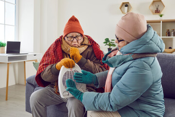 Senior family couple getting very cold at home. Two retired old people in winter coat jackets warming up by small electric fan heater, sitting together on couch in living room in cold unheated house - 796962931