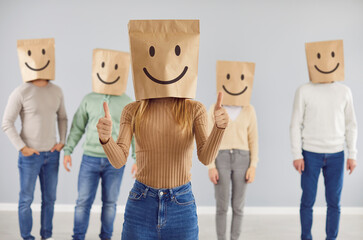 Customer with smiley bag on head feels happy, leaves positive review, gives thumbs up. Portrait of casual woman in shopping bag with cheerful face emoticon in studio with group of people in background - 796963391