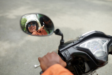 Focus on oval mirror of motorbike with reflection of young intercultural couple wearing safety helmets while moving to airport or hotel