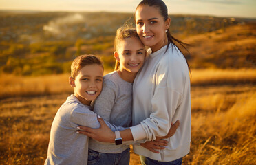 Smiling happy young mother with daughter and son standing in the field and looking at camera outdoors. Portrait of joyful woman with two children boy and girl hugging in nature. Mothers day concept. - 796966381