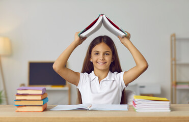 Cheerful schoolgirl who is preparing for school test in funny way, holding open book above her head. Portrait of child sitting at table among books and notebooks and having fun in front of camera. - 796969933
