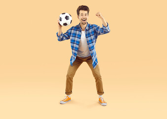 Cheerful excited man supporting his favorite soccer team during world cup match. Guy with funny facial expression holding soccer ball and clenching fist on beige background. Concept of football fans. - 796971331