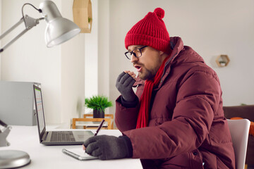 Freezing man sitting at desk blowing in his hands trying to keep warm. Warmly dressed young man in warm jacket, hat and gloves working at computer in cold winter weather. Heating problem, power crisis - 796971757