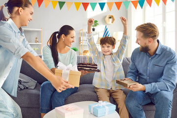 Happy Caucasian family surprises son with gifts during birthday party. He rejoices, sitting on the sofa in the living room at home, celebrating special day with happiness and the love of family. - 796973775