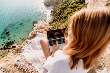 A woman is sitting on a rock overlooking the ocean with a laptop in front of her. She is enjoying the view and the peacefulness of the location.