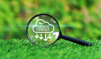 Magnifying glass focused on Carbon credit icon on the grass in garden with green nature background for reduction or removal of greenhouse gases (GHG) emissions for ecologically friendly environment.