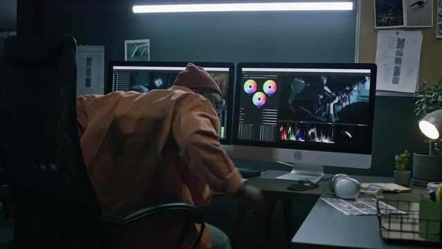 Medium rear lockdown of African American male film editor walking up to desk, sitting down in chair, putting on headphones and preparing for work on movie project in professional software