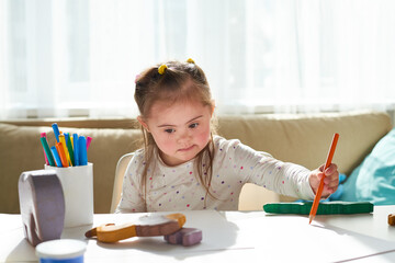 Portrait of cute little girl with Down syndrome drawing at home in sunlight, copy space
