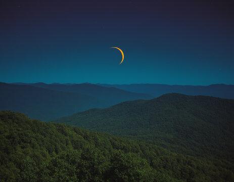 Thin Crescent Moon in a Starry Night Sky Over a Forested Landscape
