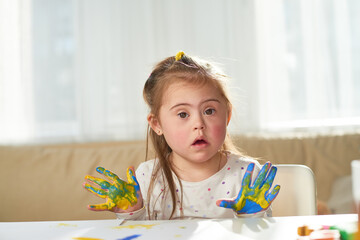 Portrait of cute little girl with Down syndrome finger painting at home in sunlight, copy space