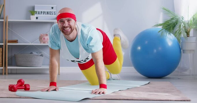 Funny nerdy Caucasian man in colorful 80s or 90s sportswear is working out and doing aerobic exercises at home. His fitness training showcases dedication to staying active and healthy.