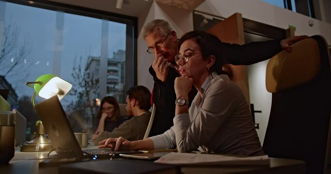 During a late evening work session, a senior male mentor advises a focused young female professional, reflecting dedication and teamwork, highlighting their commitment to mutual growth and success.