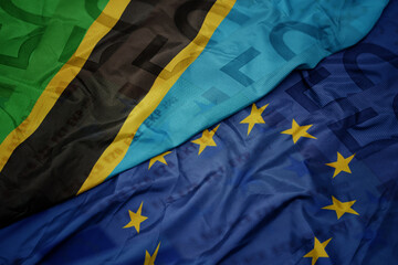 waving colorful flag of european union and flag of tanzania on a euro money banknotes background....