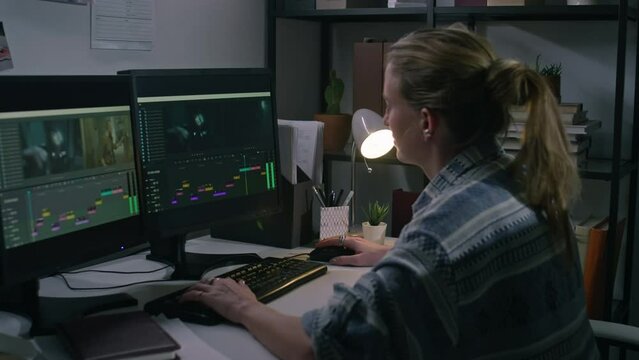 Medium side of young Caucasian female postproduction designer in casual shirt sitting at desk, editing raw footage in professional software on dual computer monitors, alone in office