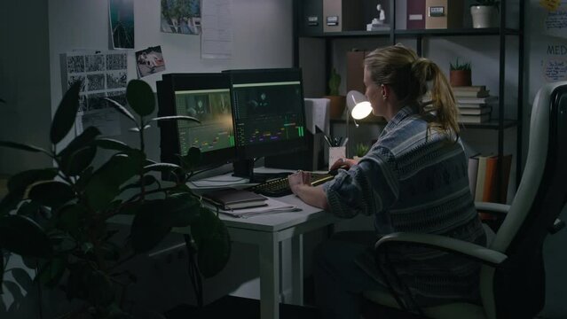 Medium full of busy Caucasian female freelance postproduction specialist in casual shirt sitting at desk at home at nighttime, editing video clips in digital software on dual monitors