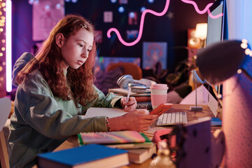 Medium shot of Caucasian girl sitting at desk in her room making notes in copybook and using...