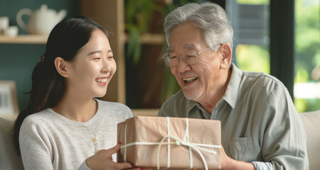 Asian adult daughter gives her senior father a gift for father's day. They smile and hug. They're sitting on the sofa at home. Concept of adult child-parent relationship, caring for single parents.