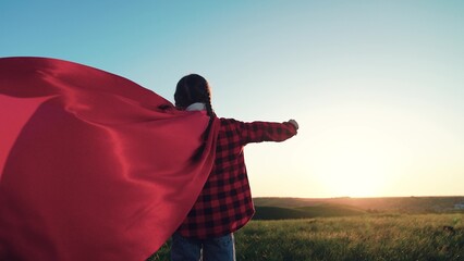 Happy child standing on green meadow wearing red superhero cape. Kid girl game concept. Child winner play in superhero costume in nature park. Little girl in red mantle play superhero against sun sky.