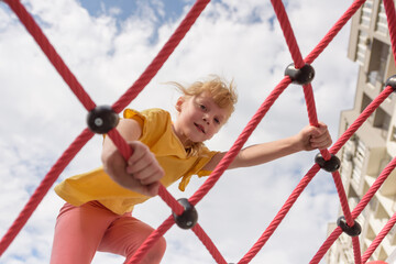 Cheerful child playing and having fun in an outdoor playground. A cute curly-haired smiling girl 6 years old in a yellow T-shirt climbs on a net. Active outdoor recreation for children in the city.