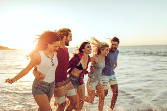 Group of friends running and laughing on the beach at sunset - Powered by Adobe