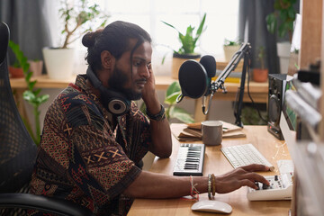 Young male zoomer in ethnic shirt sitting by desk in home studio and adjusting sound recording...