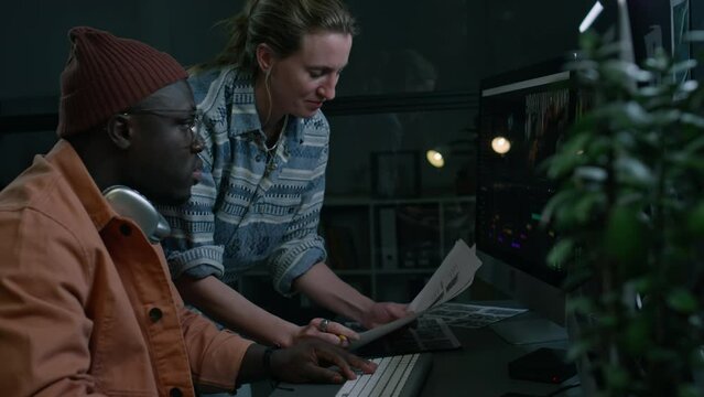 Medium side of Caucasian female producer holding movie storyboard, suggesting montage idea to African American male editor, who is working in professional software, agreeing and nodding
