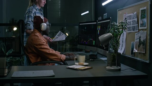 Medium fill of Caucasian female postproduction studio producer walking up to black male video editor with storyboard, discussing new ideas for frame montage, while working together on film