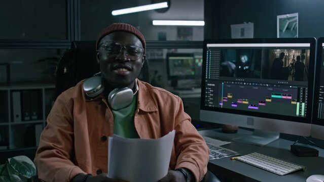 Medium portrait of cheerful young African American male video editor working on computer in professional software on raw movie footage, using script, turning on chair towards camera and smiling
