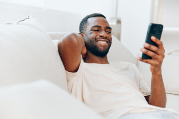 Happy African American man sitting on a black sofa, confidently typing on his mobile phone He is in his modern apartment, enjoying a moment of relaxation and connection through technology The