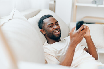 Happy African American Man Relaxing on Black Sofa, Using Smartphone and Typing Message at Home The modern apartment provides a cozy and comfortable setting for this young man as he enjoys leisure