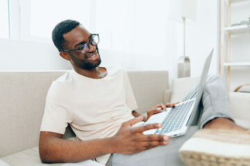 Smiling African American Man Working on Laptop in Modern Home Office, Typing and Enjoying his Freelance Job The guy is relaxed and happy, sitting on a cozy sofa in the living room The background is