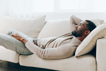 Man resting on a comfortable sofa in his modern living room, smiling and holding a laptop in his hands He is alone, enjoying a relaxing weekend at home, thinking and browsing online With a background
