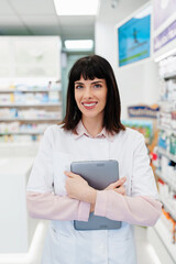 Beautiful pharmacist working and standing in a drug store and doing a stock take. Portrait of a positive healthcare worker or a chemist at his work.  She is standing and looking at camera.