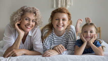 Portrait of happy older woman her young adult daughter and adorable preschooler girl smile look at camera posing lying together on bed in cozy mattress in bedroom, spend free time on weekend at home