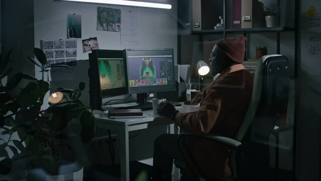 Full shot, behind glass of young African American male graphic designer in glasses, beanie sitting alone at night in studio, working in editing software on commercial footage or image, drinking coffee