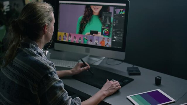 Medium rear of blonde Caucasian female freelance graphic designer sitting at desk in front of large computer monitor, editing raw commercial images, retouching with stylus pen and digital tablet