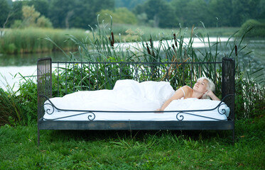 Portrait of an attractive, content, young, sexy, seductive, brunette, nude woman lying relaxed in iron bedstead bed, enjoying the nature in the autumn colors at a lake with reed and bulrush