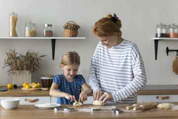 Lovely child and mom preparing buns in the kitchen. Young adult 30s mother teach her adorable preschooler 6s daughter to cook, making together homemade dough, smiling, enjoy dessert cooking at home