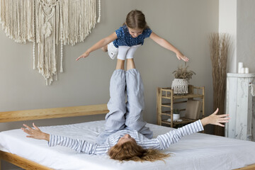 Little girl pretends flying in air enjoy playtime with mother lying on bed lifts her cute preschooler daughter up on outstretched legs, exercising, spend active time in cozy bedroom on weekend at home