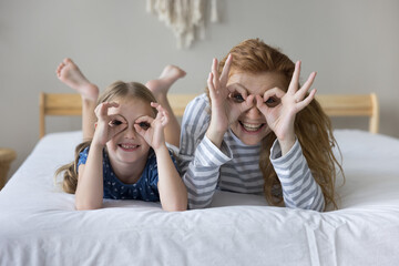 Happy little playful daughter and young mother lying on bed looking at camera through fingers, make circles, binoculars or eyewear shape. Family having fun, advertising vision check-up, eyesight care