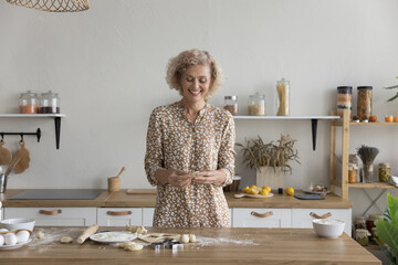 Attractive middle-aged woman in elegant casual dress standing alone in modern kitchen, smiling,...
