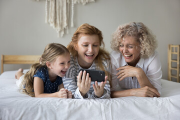 Happy little girl spend time with mum and grandmother at home, lying in bed with smart phone, laughing while watch funny videos in social networks, have fun using new application, enjoy internet usage